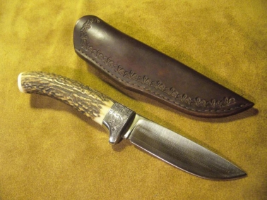 Vad&aacute;szk&eacute;s szarvasagancs markolattal, rozsdamentes ac&eacute;l k&eacute;zv&eacute;dővel &eacute;s San Mai damaszkpeng&eacute;vel. A cizell&aacute;l&aacute;s &Aacute;rva Edmund fegyverv&eacute;sn&ouml;k munk&aacute;ja.